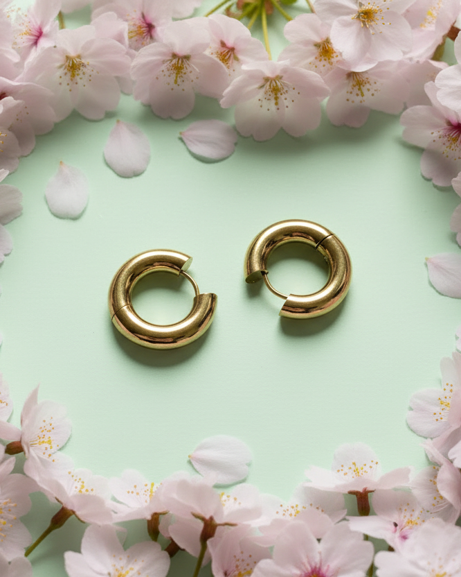 Zwei goldene, Tube-Creolen liegen auf einem hellgrünen Hintergrund, eingerahmt von Kirschblüten und Blütenblättern.