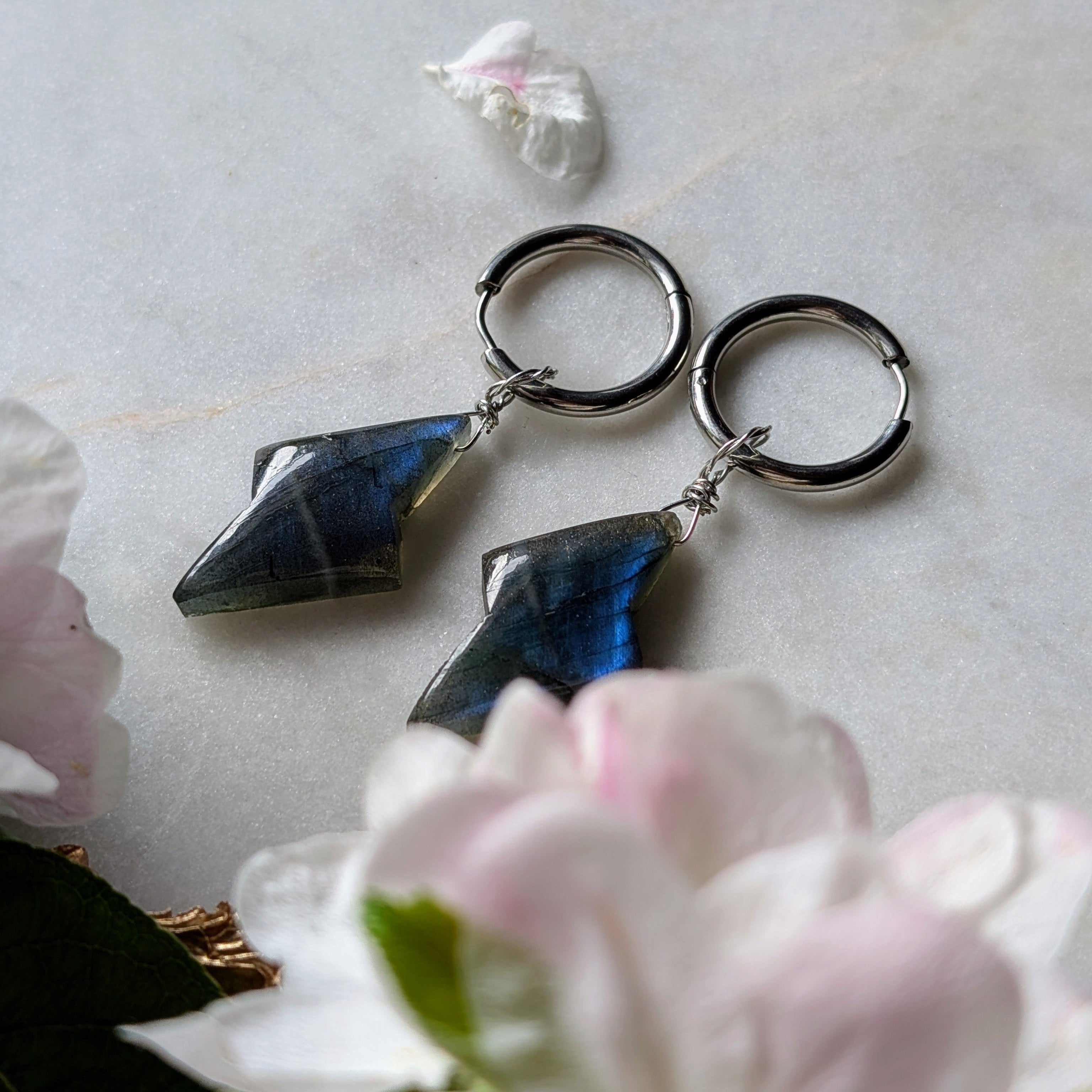  Edelstahl Creolen mit Anhängern aus blauem Labradorit geformt wie ein Blitz. Alles liegt auf einem Marmorteller, dekoriert mit Apfelblüten 