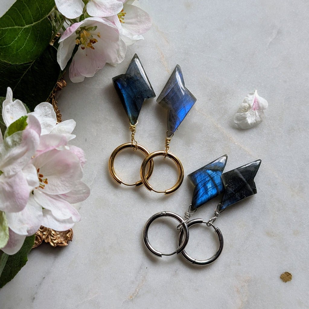 goldene und silberne Edelstahl Creolen mit Anhängern aus blauem Labradorit geformt wie ein Blitz. Alles liegt auf einem Marmorteller, dekoriert mit Apfelblüten 