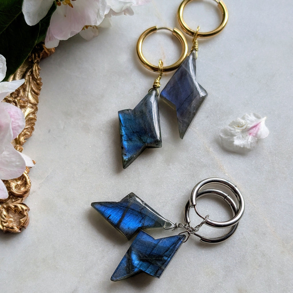 goldene und silberene Edelstahl Creolen mit Anhängern aus blauem Labradorit geformt wie ein Blitz. Alles liegt auf einem Marmorteller, dekoriert mit Apfelblüten 