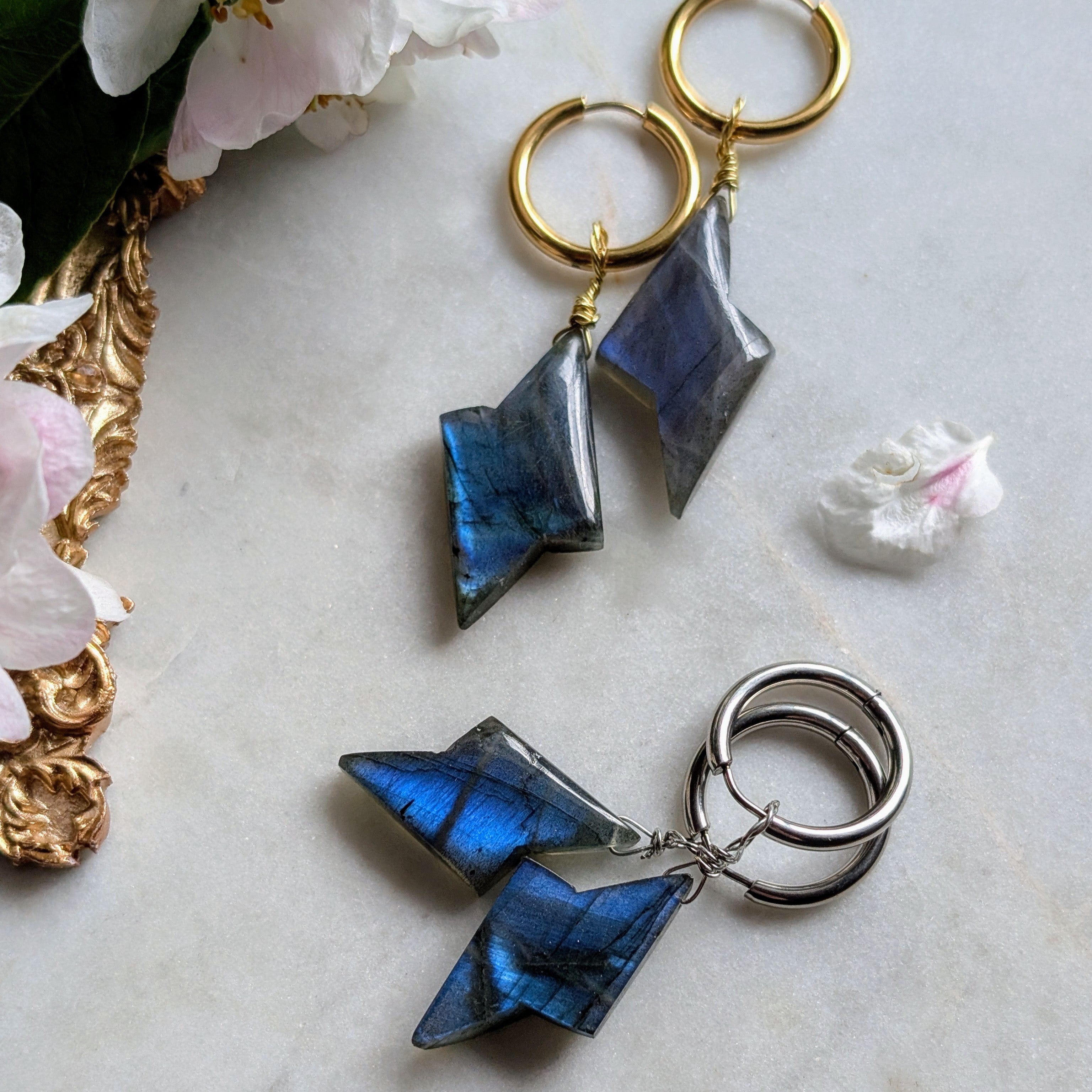 goldene und silberene Edelstahl Creolen mit Anhängern aus blauem Labradorit geformt wie ein Blitz. Alles liegt auf einem Marmorteller, dekoriert mit Apfelblüten 