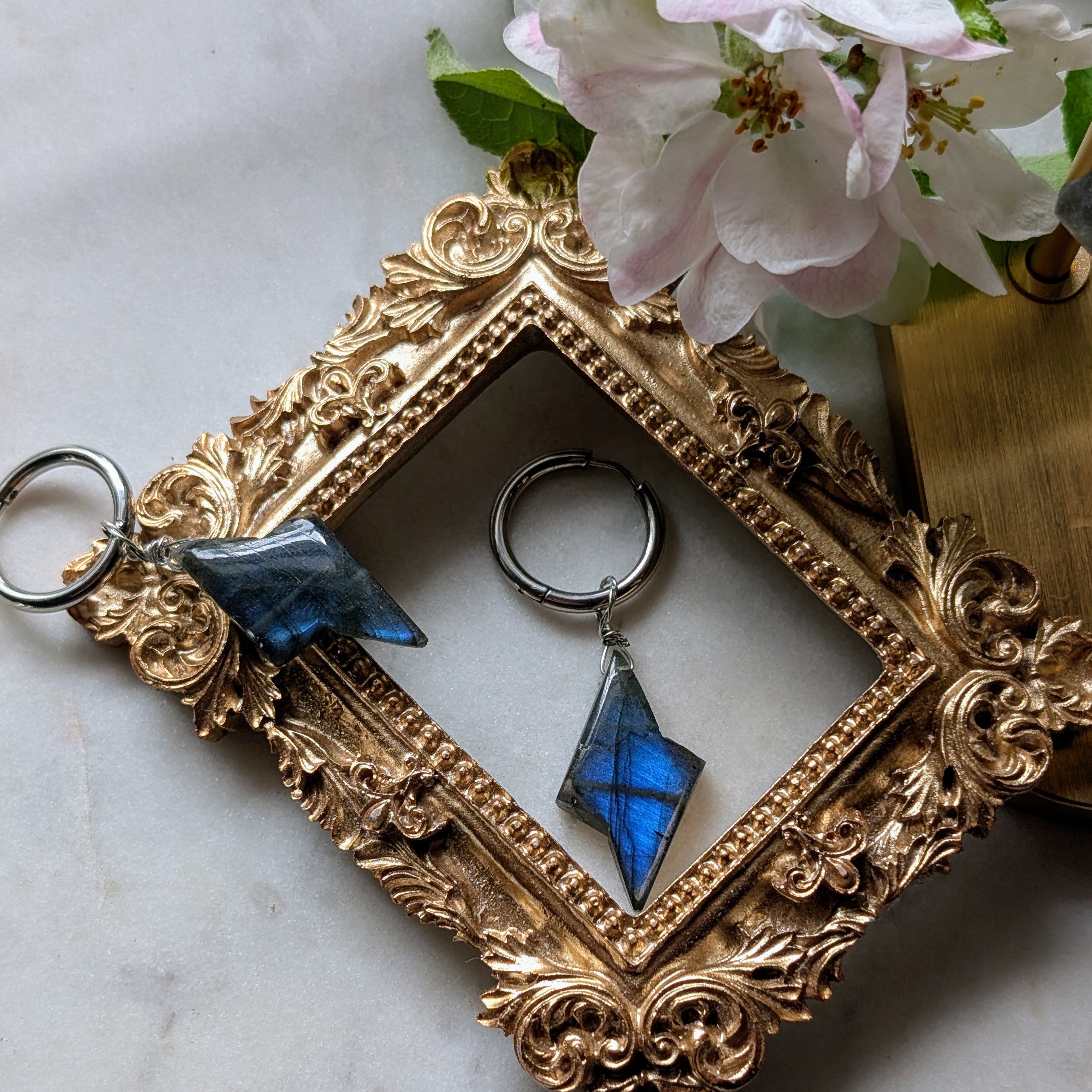 Edelstahl Creolen mit Anhängern aus blauem Labradorit geformt wie ein Blitz. Alles liegt auf einem Marmorteller, dekoriert mit Apfelblüten 