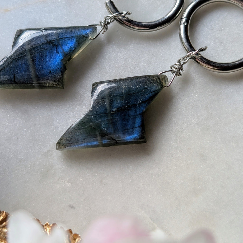 Edelstahl Creolen mit Anhängern aus blauem Labradorit geformt wie ein Blitz. Alles liegt auf einem Marmorteller, dekoriert mit Apfelblüten 