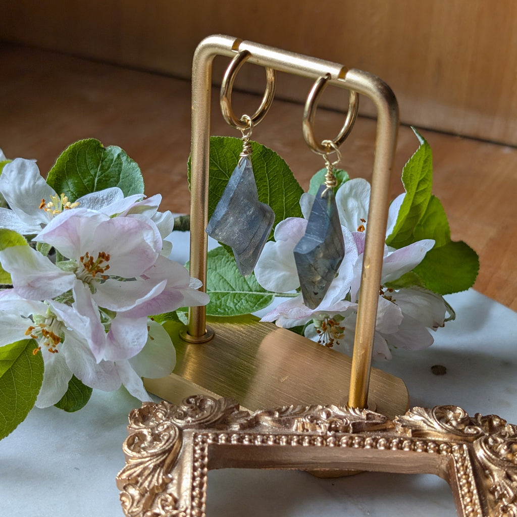 goldene Edelstahl Creolen mit Anhängern aus blauem Labradorit geformt wie ein Blitz. Alles liegt auf einem Marmorteller, dekoriert mit Apfelblüten 