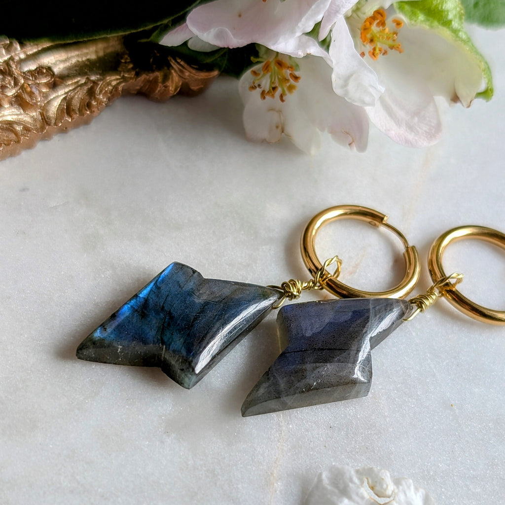 goldene Edelstahl Creolen mit Anhängern aus blauem Labradorit geformt wie ein Blitz. Alles liegt auf einem Marmorteller, dekoriert mit Apfelblüten 