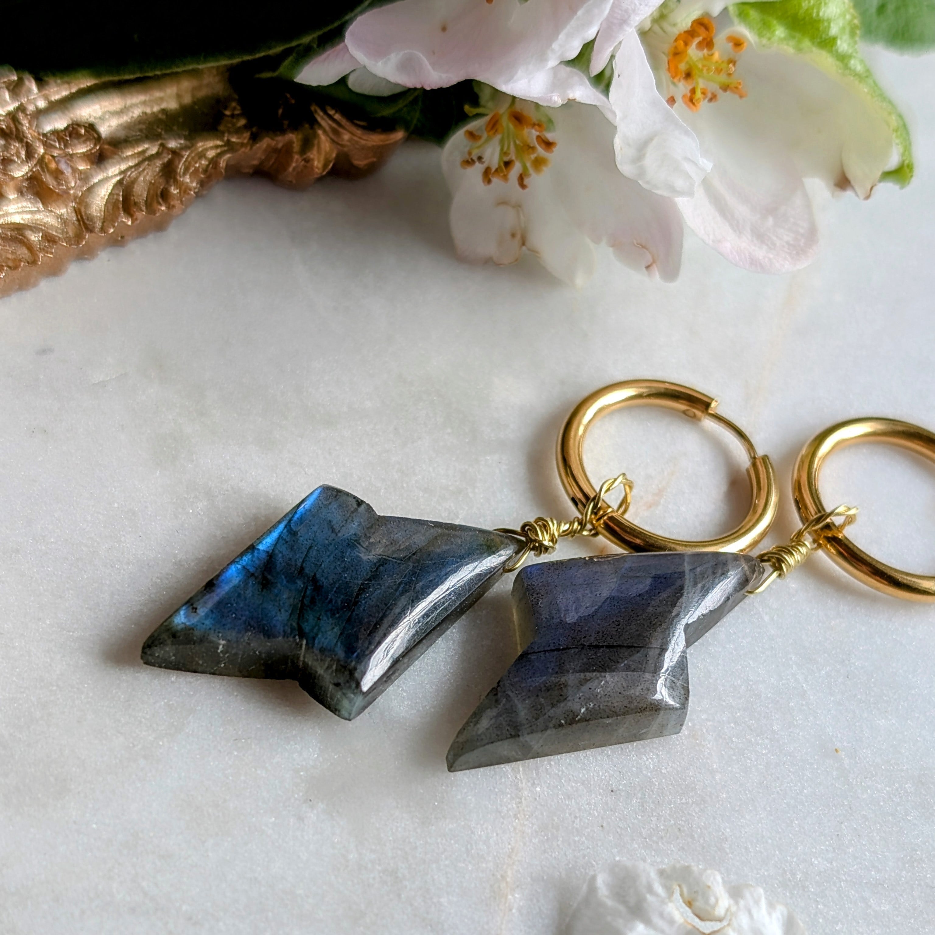 goldene Edelstahl Creolen mit Anhängern aus blauem Labradorit geformt wie ein Blitz. Alles liegt auf einem Marmorteller, dekoriert mit Apfelblüten 