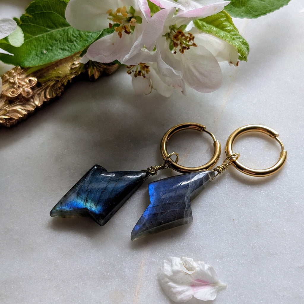goldene Edelstahl Creolen mit Anhängern aus blauem Labradorit geformt wie ein Blitz. Alles liegt auf einem Marmorteller, dekoriert mit Apfelblüten 