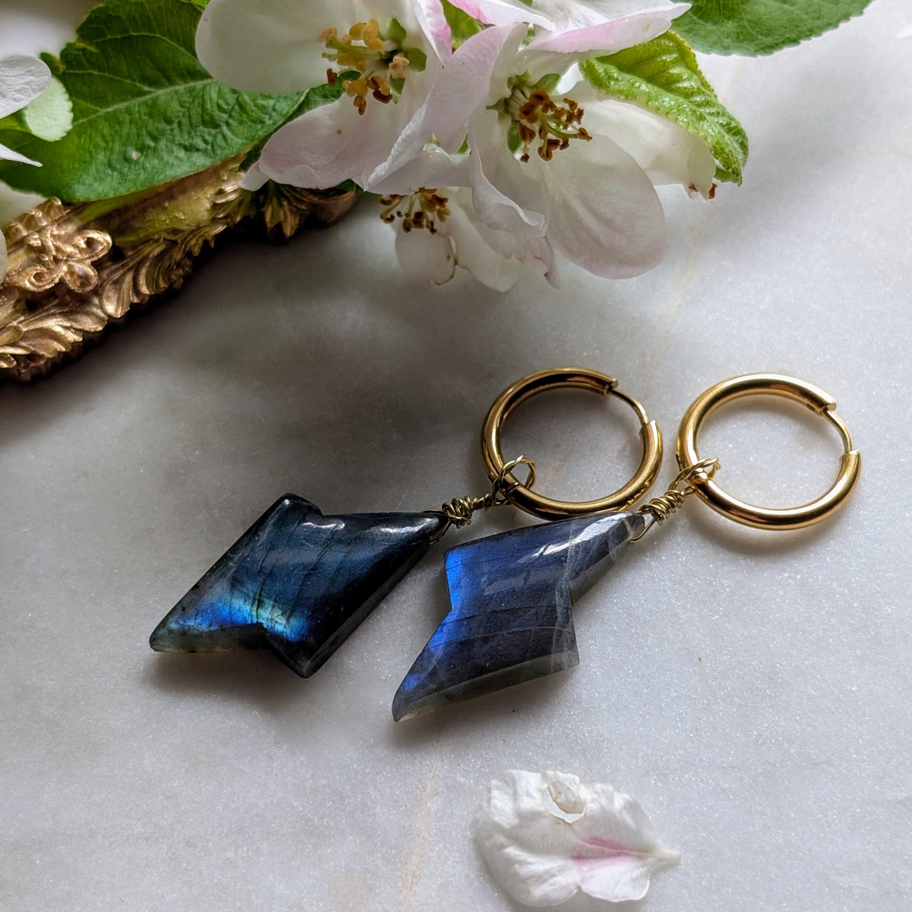 goldene Edelstahl Creolen mit Anhängern aus blauem Labradorit geformt wie ein Blitz. Alles liegt auf einem Marmorteller, dekoriert mit Apfelblüten 