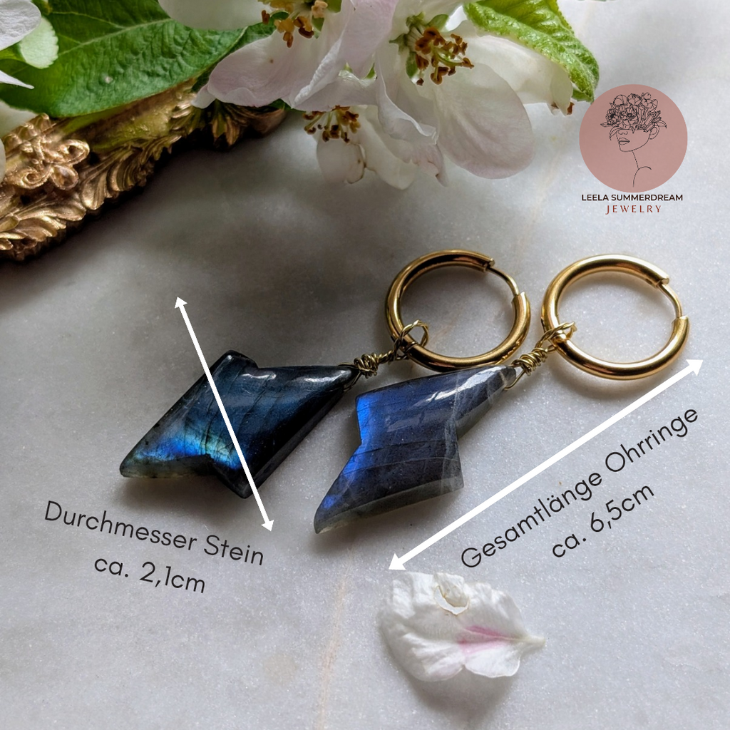 goldene Edelstahl Creolen mit Anhängern aus blauem Labradorit geformt wie ein Blitz. Alles liegt auf einem Marmorteller, dekoriert mit Apfelblüten 