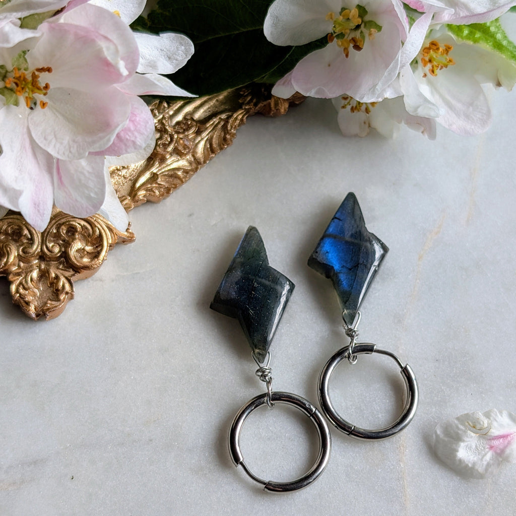 Edelstahl Creolen mit Anhängern aus blauem Labradorit geformt wie ein Blitz. Alles liegt auf einem Marmorteller, dekoriert mit Apfelblüten 