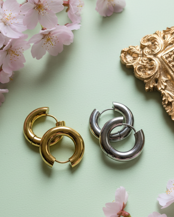 Nahaufnahme von silbernen und goldenen „Chunky“ Creolen, die zwischen frischen Kirschblütenzweigen auf einem grünen Untergrund präsentiert werden.