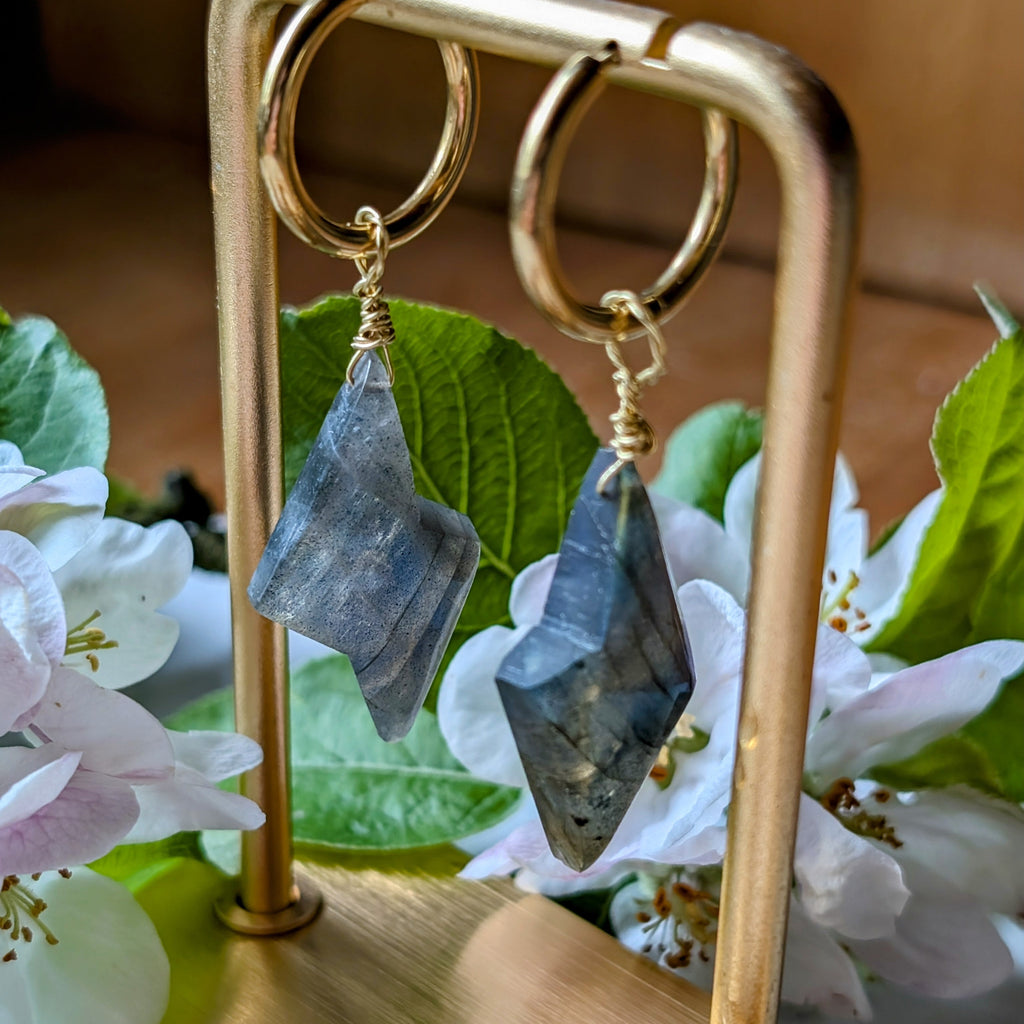 goldene Edelstahl Creolen mit Anhängern aus blauem Labradorit geformt wie ein Blitz. Alles liegt auf einem Marmorteller, dekoriert mit Apfelblüten 