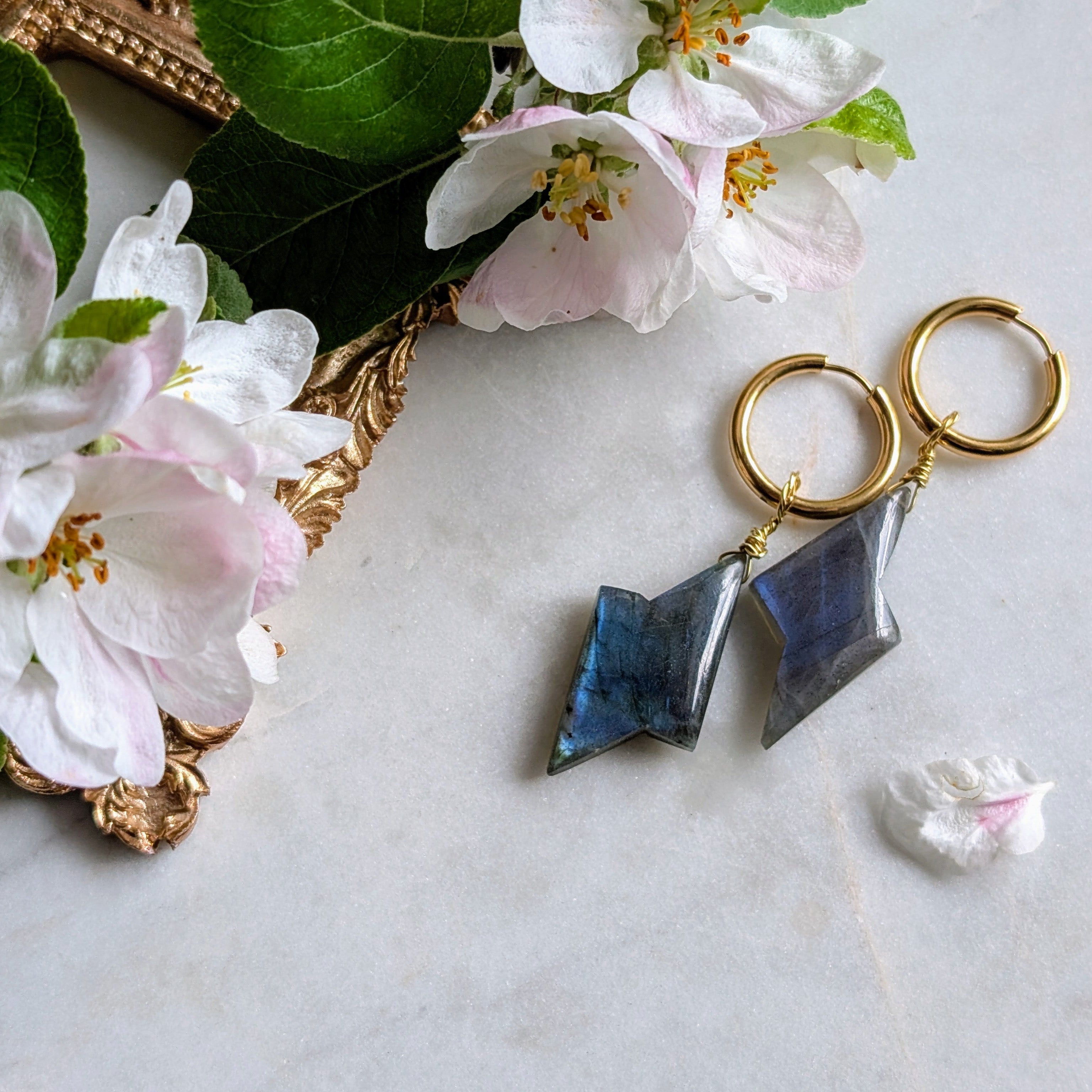 goldene Edelstahl Creolen mit Anhängern aus blauem Labradorit geformt wie ein Blitz. Alles liegt auf einem Marmorteller, dekoriert mit Apfelblüten 