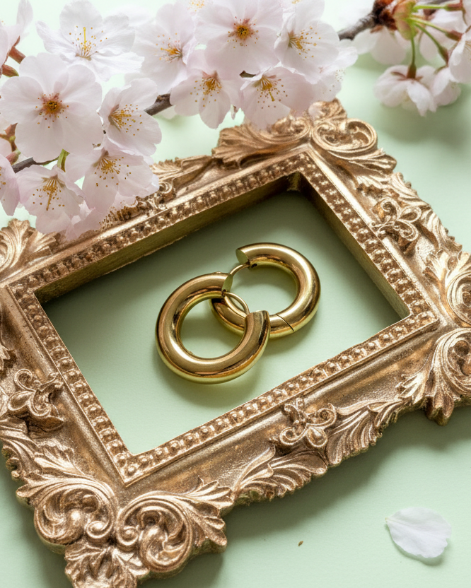 Ein goldener Barockrahmen auf mintgrünem Untergrund umschließt ein Paar goldene „Chunky“ Creolen, dekoriert mit einzelnen Kirschblütenblättern