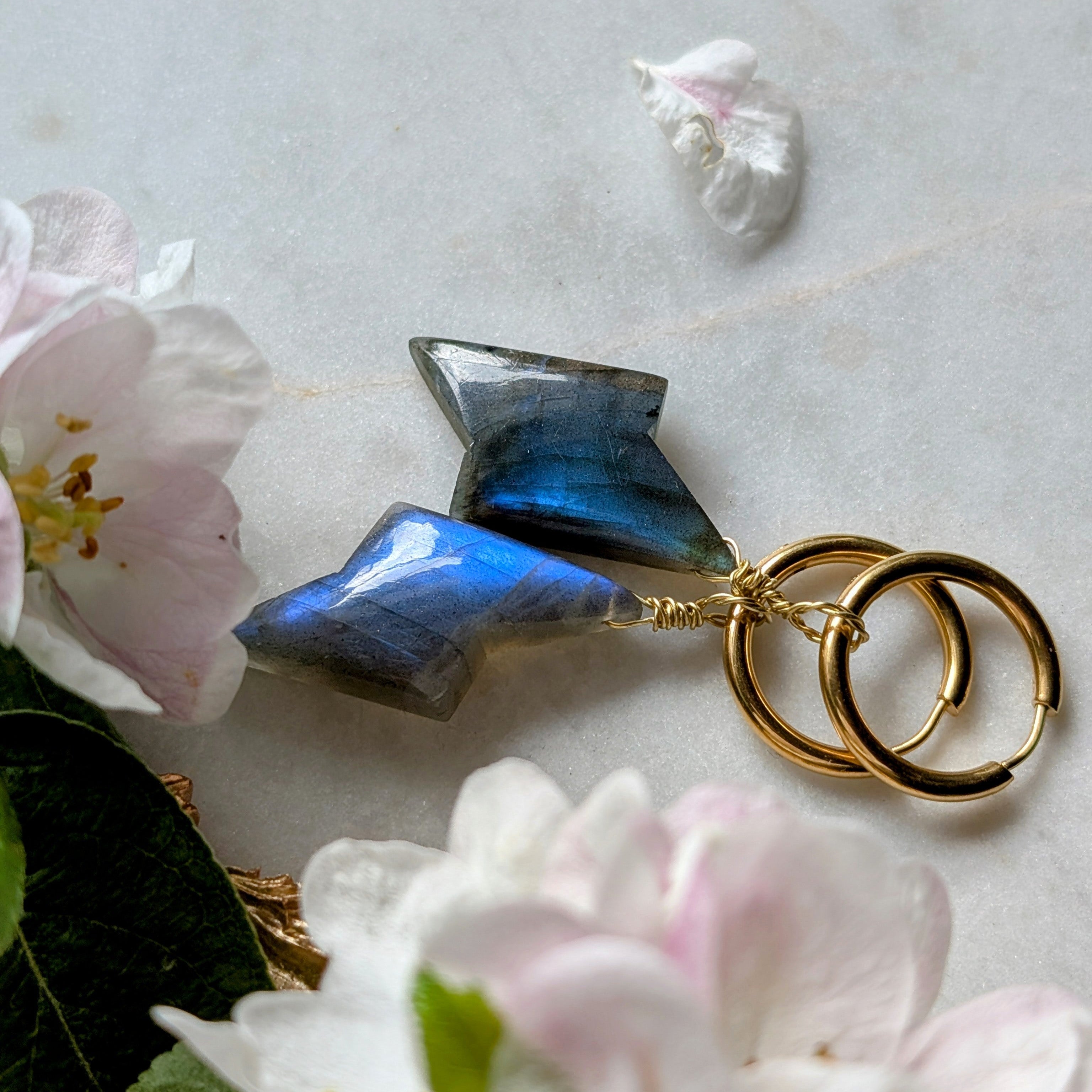 goldene Edelstahl Creolen mit Anhängern aus blauem Labradorit geformt wie ein Blitz. Alles liegt auf einem Marmorteller, dekoriert mit Apfelblüten 