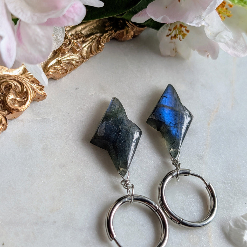 Edelstahl Creolen mit Anhängern aus blauem Labradorit geformt wie ein Blitz. Alles liegt auf einem Marmorteller, dekoriert mit Apfelblüten 